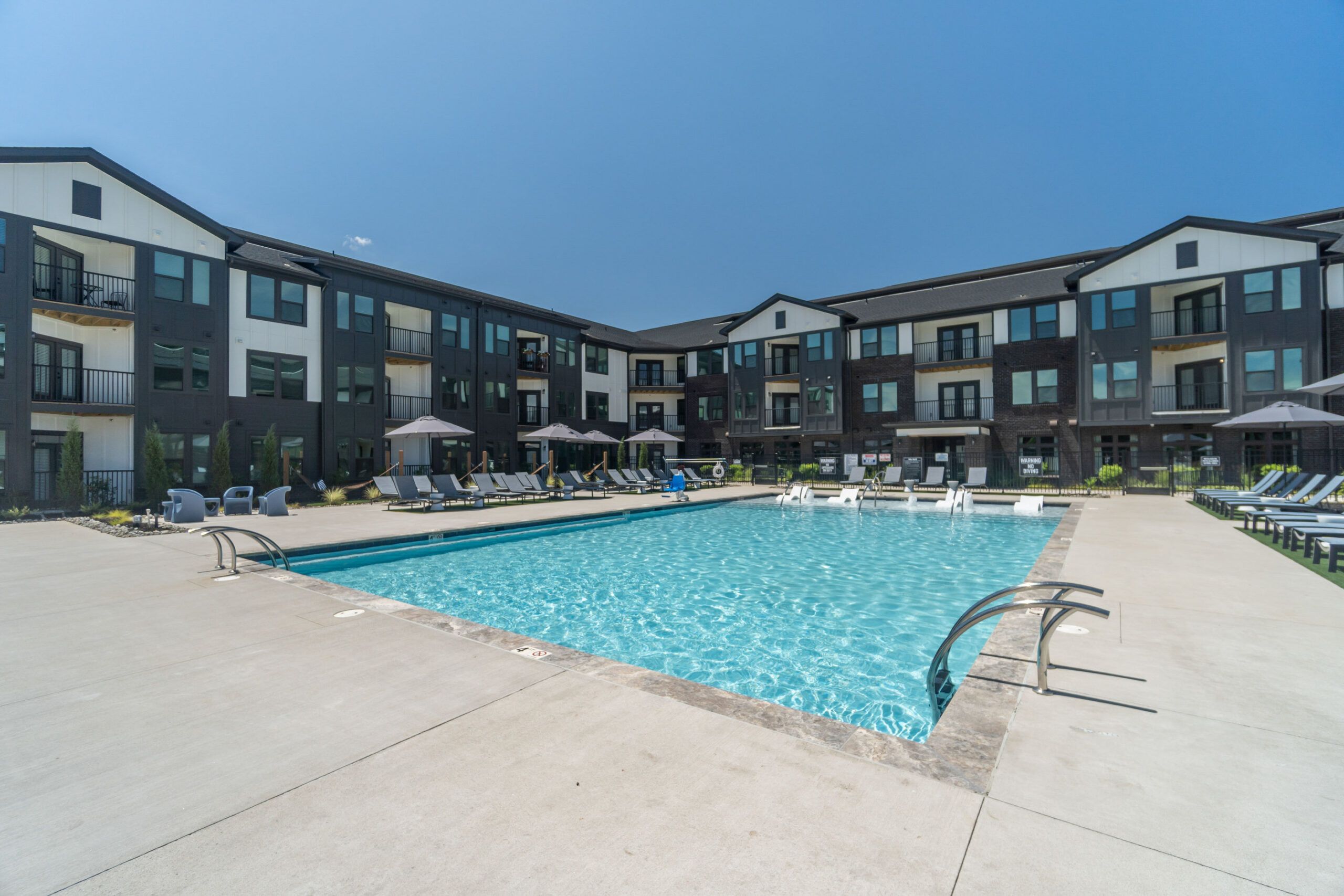 A sparkling, resort-style swimming pool at Evolve at Lake Norman surrounded by a spacious concrete deck and modern apartment buildings under a clear blue sky.